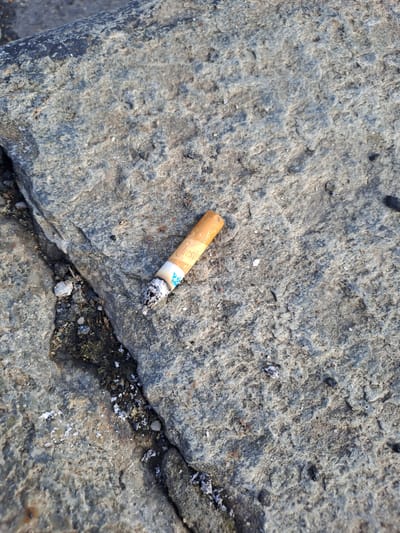 The image shows a close-up, top-down view of a discarded cigarette butt resting on a rough, grey stone surface. The cigarette is partially burned, with ash at the tip. The stone has visible textures and cracks, with some debris and small white particles scattered on its surface. The lighting suggests an outdoor environment, possibly in a public space. There is no visible indication of the specific location within the city of Turin, Italy, or any context related to people or actions.