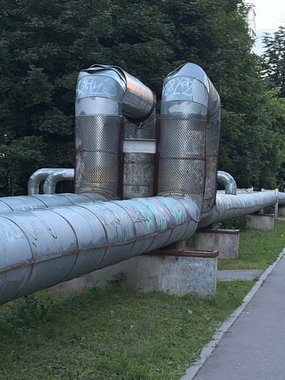 The image shows a section of large, insulated pipes, likely for district heating or industrial use, running horizontally across the frame from right to left. The pipes are metallic gray with some signs of wear, including visible seams and patches of light green or turquoise coloring, possibly from weathered paint or corrosion. Some sections are covered with a wire mesh. White graffiti is present on some of the pipes, with illegible tags.
The pipes are supported by concrete blocks arranged along their length. To the left of the pipes, a dense cluster of dark green trees forms a natural backdrop, suggesting an outdoor setting. To the right, there is a strip of green grass adjacent to a paved surface, which could be a sidewalk or a road. The lighting suggests it is daytime, possibly late afternoon or early evening, given the soft light and shadows. There are no people visible in the image. The overall impression is of urban infrastructure integrated into a partially natural environment. The location context is Moskovskiy, Russia.