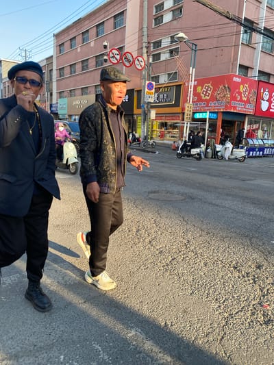 Two men stand on a paved street in Jinzhou, China. The man in the foreground wears sunglasses and a dark jacket, possibly holding a cigarette. The second man is older, wearing a cap and patterned jacket. Behind them is a street with shops displaying Chinese characters. Signs include a reference to "JIAREN YUAN" and "PASTIY" indicating a bakery. Another sign advertises grilled seafood. Several parked scooters line the street. Traffic signs are also visible. Buildings with multiple windows form the backdrop.