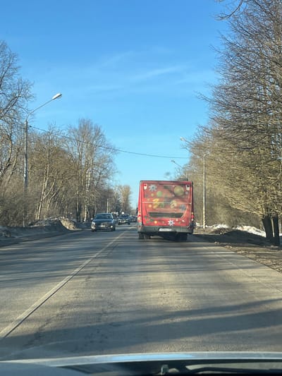 An image captured from the interior of a vehicle shows a multi-lane asphalt road in Yasenevo, Russia, under a clear blue sky. Ahead, a red public transit bus with "МОСТРАНСАВТО" printed on its rear, featuring colorful graphics depicting planets, stars, flowers, and Easter eggs, travels in the right lane. Its Russian license plate is H632XX 199. Several other passenger vehicles, including a dark-colored sedan, are visible further ahead and to the left of the bus, proceeding in the same direction. The road surface appears worn in places, with a solid white line separating the two lanes. Bare deciduous trees line both sides of the road, suggesting a late winter or early spring season. Piles of residual snow and dirt are present along the road shoulders. Multiple streetlights with horizontal fixtures are positioned along the left side of the thoroughfare. The scene is illuminated by natural daylight, casting shadows on the road.