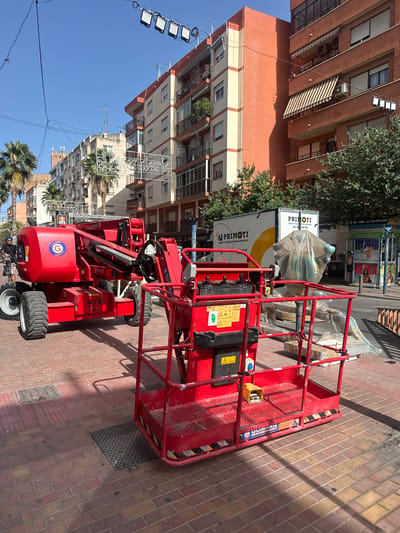 This is a medium shot of a red aerial work platform parked on a brick sidewalk in a city. The platform is the primary subject, with its metallic cage and hydraulic arm visible. The background features buildings with apartment balconies and awnings, suggesting an urban residential area. Trees and palm trees are also present, indicating a warm climate. A white truck with "PRIMOTI" and "alquiler y renting de vehiculos industriales" written on it is parked behind the platform. The sun is high in the sky, casting shadows and suggesting it is daytime, likely mid-morning or early afternoon. The sky is clear and blue. Visible text on the platform includes "GOMARIZ" and "4x4," along with various safety symbols and a circular logo with a "G" inside. A person wearing a helmet and riding a bicycle is visible in the lower left corner. The scene appears to be a street in Alicante, Spain, possibly during a construction or maintenance job.