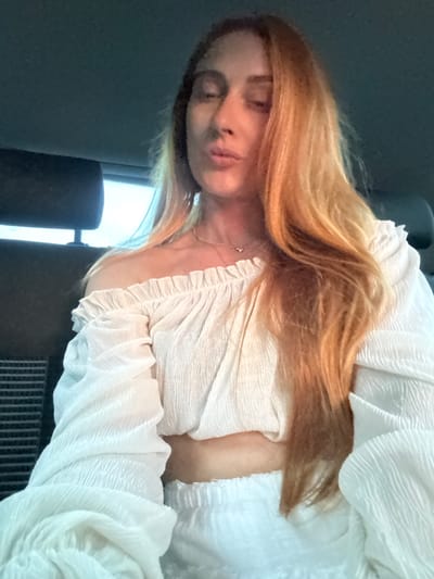 The scene is set inside a vehicle in Podličak, Montenegro. A woman with long, flowing reddish-blonde hair occupies the majority of the frame. Her eyes are closed or heavily lidded, and her lips are pursed in a slight pucker. Her complexion appears fair. She wears a white, off-the-shoulder crop top featuring a ruffled elastic neckline and textured, possibly crinkle-fabric, long sleeves. A segment of her bare midriff is visible above white lower garments, likely shorts or a skirt, made of similar textured fabric. A thin, light-colored necklace with a small embellishment is visible around her neck.
The interior of the vehicle is dimly lit, predominantly dark grey or black. A dark headrest from a car seat is visible behind her, partially obscuring a bright, rectangular light source (likely a window) in the background on the left. Another part of the car's interior, possibly an air vent or grating, is visible in the lower-left corner of the frame. The ceiling of the vehicle, also dark, is visible in the upper portion of the image. Strong directional light, possibly from an external window, illuminates the right side of her face and hair, creating highlights, while the left side of her face is in softer shadow.