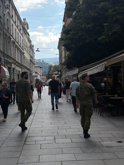 The image depicts a street scene in Sarajevo, Bosnia and Herzegovina, specifically in the Baščaršija district, a historic old town. In the foreground, two individuals in camouflage military uniforms are walking away from the camera on a cobblestone street lined with buildings and outdoor seating for cafes. The man on the left is wearing a green and brown camouflage uniform with a black belt and black boots. He is looking slightly over his left shoulder. The woman on the right is also in a camouflage uniform with her hair in a braid. She is looking straight ahead.
Several other pedestrians are visible further down the street, also walking in various directions. The buildings on either side of the street are tall and have a European architectural style, with multiple stories and windows. On the right side, there are awnings extending from cafes and shops, with tables and chairs set up outside. The sky above is partly cloudy. In the background, a hilly landscape with buildings can be seen. The overall atmosphere appears to be a busy daytime scene in an urban environment.