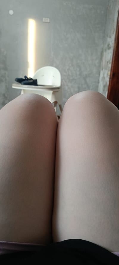 The image shows a close-up view of a person's thighs. In the background, a white high chair with black shoes on it is visible, along with a wall with an electrical outlet and a window with light shining through it. The setting appears to be indoors. The image is not specific enough to determine the location in the city of Хохлы, Russia.