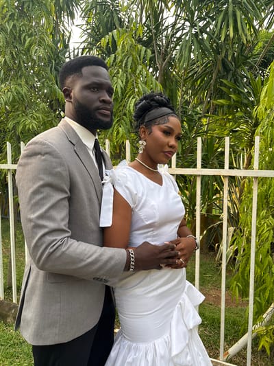 This image depicts a couple posing outdoors, likely for a wedding or engagement photoshoot, in Barikin Gidar Kanat, Nigeria. The woman is wearing a white dress with a pearl necklace and elaborate earrings. Her hair is styled in braids, and she has dramatic eye makeup. The man is dressed in a grey suit with a white shirt and black tie. They are holding hands, with the woman's left hand resting on the man's arm and his hand gently holding her abdomen. A ring is visible on the woman's left hand. The background consists of lush green foliage, suggesting a garden or natural setting. A white metal fence is also visible, partially obscured by the greenery. The lighting indicates it is daytime, likely with bright sunshine. The couple's poses and gazes suggest a romantic and celebratory mood.