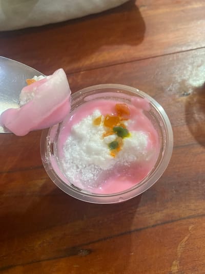 A close-up, overhead shot shows a spoon taking a bite of a dessert in a clear plastic cup. The dessert is a pink, creamy substance with a scoop of white, shaved ice or coconut in the center. It is garnished with small, colorful pieces of fruit, appearing to be orange and green. The dessert sits on a dark, wooden surface, likely a table. The background is blurred, showing hints of a light-colored fabric. The location in Ta', Indonesia is not discernible from the image.