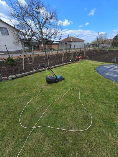 The image shows a garden scene in Stara Reka, Bulgaria. A green lawn is the primary subject, with a dark grey and green lawnmower positioned on it. A long, white electrical cord extends from the lawnmower across the grass. Adjacent to the lawn is a garden plot with exposed dark soil and a fence line. A building is visible in the background, partially obscured by a bare tree. The sky is blue with scattered white clouds.