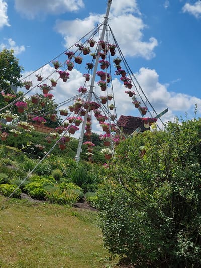 The image shows an outdoor scene in Kovalivka, Ukraine, featuring a large, conical-shaped structure adorned with numerous hanging potted flowers. This structure, possibly a decorative art installation or a unique garden feature, is supported by a central pole and appears to have tiered, angled elements from which the flower pots are suspended by chains. The pots themselves are small and brown, containing pink, white, and possibly purple petunias.

The background consists of a bright blue sky with scattered white clouds. To the left of the central structure, there is a lush green tree. The ground below is covered with various types of vegetation, including a manicured lawn in the foreground, and meticulously planted flower beds and shrubs with various shades of green. In the middle ground, to the right of the hanging flower structure, a large, dense green bush dominates the right side of the frame, partially obscuring the view of the ground and some of the vegetation behind it. A hint of a windmill-like structure covered in red flowers is visible behind and to the right of the hanging flower installation. The scene is well-lit, suggesting it was taken on a sunny day. There are no people visible in the image.