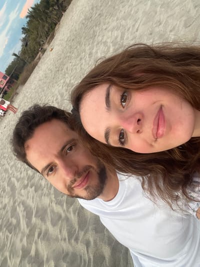 A man and a woman pose for a selfie on the beach in Brisas de Zicatela, Mexico. The woman is wearing a floral shirt and has brown hair. The man is wearing a white t-shirt and has dark hair and a beard. The background shows palm trees, a pink building, and other people on the beach.
