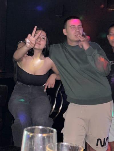 The image depicts three individuals in a dimly lit indoor establishment, likely a bar or club, located in Lom, Bulgaria. In the foreground, two clear drinking glasses are visible, out of focus.
On the left, a young adult female is seated. She has dark hair and is wearing a black strapless top, dark grey jeans, and a delicate silver-colored necklace with a small pendant. Her fingernails are painted white. She holds her left hand up, displaying a "peace" sign (V-sign with index and middle fingers). Her right arm rests on the armrest of her chair or the man's arm. She looks directly towards the camera with a slight smile. A distinct yellow light is reflected on her neck and chest, and blue and green lights are reflected on her jeans.
To her right, a young adult male stands, with his left arm around her shoulders. He has short, dark hair and is wearing a dark green long-sleeved shirt and light beige shorts featuring a black "NASA" logo on the left leg. He holds a cigarette or vape pen to his mouth with his right hand. He looks towards the camera, his expression neutral. A pink light is reflected on his head and face, and an orange-red light is visible on his right sleeve.
Partially visible on the far right is a third individual, likely a young adult male, with only a portion of his face and shoulder showing. An orange light reflects on his cheek. The background is dark and out of focus, with various colored lights (pink, green, blue, yellow, red) indicating disco or party lighting. The overall scene suggests a social gathering in an entertainment venue.