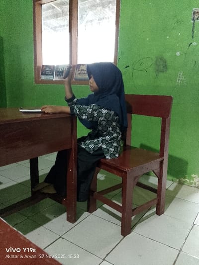 A young girl is seated at a dark brown wooden desk and chair in a room in Wanguk, Indonesia. She is viewed in profile, facing left, wearing a dark blue hijab, a long-sleeved black shirt with a white and green floral/geometric pattern, and dark pants. Her left hand rests on the desk near a white paper or book. Her right hand is raised, holding a dark object (possibly a mobile phone) to her ear. The room features bright green walls with some worn patches and dark markings, including a circular doodle. A rectangular window with a dark frame is visible behind her, revealing bright light and a corrugated roof structure outside. Several small, dark objects resembling books or frames are visible on a ledge beneath the window. The floor is covered with light-colored square tiles. Shadows are cast on the floor beneath the furniture. The image was captured by a vivo Y19s device on November 27, 2025, at 15:23.