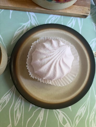 A single pink marshmallow sits in a fluted white paper liner on a brown-rimmed plate. The plate rests on a green placemat with a white leaf pattern. The scene is viewed from directly above.