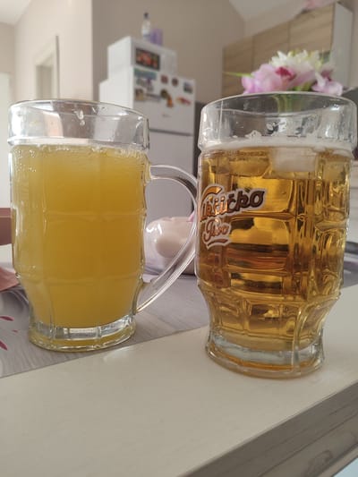 Two glasses are positioned side-by-side on a white wooden surface. The glass on the left is filled with a bright yellow, bubbly liquid, possibly juice or soda. It has a handle. The glass on the right contains a golden-amber colored liquid, likely beer, with ice cubes and a frothy head. This glass also has a handle and features a logo with text that appears to read "Likicko pivo". In the background, a refrigerator, kitchen cabinets, and a bouquet of flowers are visible. The scene is indoors, suggesting a domestic or restaurant setting. The exact location within Škaljari, Montenegro cannot be determined from the image.