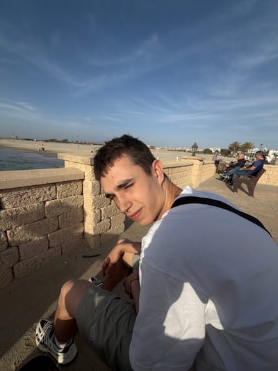 A medium-shot photograph taken outdoors, rotated clockwise, depicts a young man on a paved area with a brick wall next to a beach. The man has short, dark hair and is wearing a white t-shirt and gray shorts with a logo of "The North Face" on his left thigh. He is also wearing white sneakers. He is leaning towards the camera with his head near the brick wall. In the background, there is a beach with water and people sitting on benches under palm trees. The sky is visible above the scene and is blue with some clouds. The photograph was taken in Kiryat Yam, Israel.