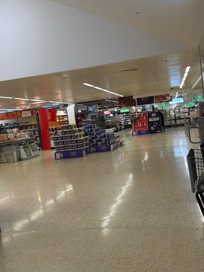 This is an interior shot of a supermarket aisle in Pity Me, United Kingdom. The aisle is long and well-lit with overhead fluorescent lights. The floor is a textured beige tile. Merchandise is arranged on shelves and in displays along both sides of the aisle. Brands visible include Pepsi, Fosters, and Nectar. The aisle also has signage related to 'Made for summer' products. There is a shopping trolley at the very top of the aisle. The angle suggests the photo was taken while standing still near the beginning of the aisle.
