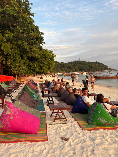 The image captures a beach scene at sunset or golden hour in Ban Ko Li Pe, Thailand. The sky exhibits a gradient of light blue with scattered, wispy white clouds, illuminated by warm, low-angle sunlight.
In the foreground, a white sandy beach hosts multiple rows of beach seating arrangements. Each setup consists of a brightly colored, patterned beanbag chair placed on a striped woven mat, accompanied by a small, dark wooden table. Beanbag colors include pink, green, blue, and various multi-patterned designs, while mats feature green, red, and yellow stripes. Several tables contain small ashtrays.
Multiple individuals are visible, primarily seated or reclining on the beanbags. In the immediate foreground, a person is seated on a bright green beanbag, and another on a blue beanbag. Further back, a man in a white t-shirt is seated on a blue beanbag. Other patrons are scattered among the seating, some conversing, others facing towards the water.
Along the sandy shoreline, numerous people are walking or standing. A woman in a bikini top and shorts is seen walking towards the right, and a man in shorts stands near the water's edge. Additional groups of individuals populate the distant beach area.
The calm, light blue-green sea in the mid-ground contains several traditional Thai longtail boats and smaller motorboats, either anchored or moored. To the far left, a dense canopy of large green trees borders the beach. In the background, a heavily forested island or hill rises from the water, topped by a distinct communication tower or antenna structure.