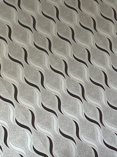 This media file presents a detailed, close-up view of a decorative patterned surface, likely wallpaper or a similar interior finish, located in Zetiovo, Bulgaria. The primary subject is an abstract, repeating pattern composed of flowing, curvilinear wave-like shapes.
The design features a sophisticated monochromatic palette, predominantly in shades of gray, white, and a deep brown or almost black. The background areas of the pattern are rendered in a lighter gray with a noticeable speckled or granular texture, providing a tactile feel. Layered upon this background are multiple parallel wavy lines. Some waves are a pristine white, while others are a medium gray, creating an illusion of depth and movement. Interspersed between these lighter waves are bold, solid dark brown/black serpentine lines that sharply define the pattern and add visual weight.
The wavy elements are arranged diagonally across the entire image, forming an interlocking grid that creates a dynamic and rhythmic visual flow. There are no people, objects, or specific scenes depicted beyond this decorative surface. No activity or event is taking place. The image does not provide cues regarding the time of day, weather conditions, or any emotional context. There is no visible text present in the image. The pattern conveys a modern and elegant aesthetic, characteristic of contemporary interior design.