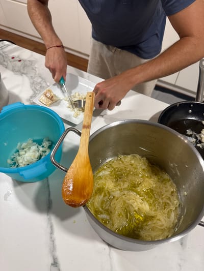 The scene depicts a kitchen in Cidade da Maia, Portugal. A person, visible from the mid-torso down, wears a blue t-shirt and light-colored shorts. Their arms are positioned over a white marble-patterned countertop. The person is actively using a knife with a light blue handle in their right hand and a silver fork in their left hand to flake or chop white fish on a rectangular white plate. A piece of cooked fish remains on the plate. To the left, a light blue plastic bowl contains a quantity of flaked white fish. A large silver pot on the counter contains finely sliced, translucent onions simmering in a clear liquid, consistent with olive oil. A wooden spoon, coated in the liquid and featuring a small hole near its top, rests diagonally on the rim of this pot. To the far right, a dark frying pan contains small, browned fragments, possibly garlic or fish, in residual oil. White cabinets are visible in the background above a dark floor.