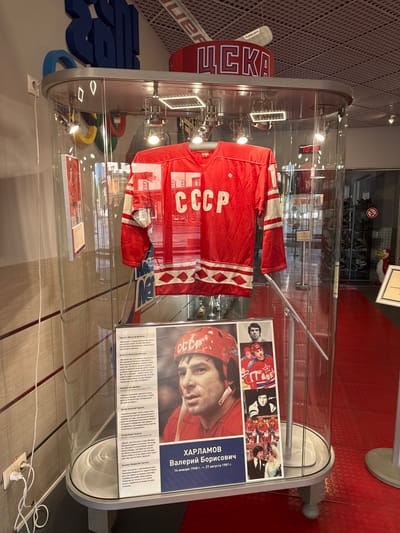 The image depicts a display case in Peresvet, Russia, containing a Soviet Union ice hockey jersey, a hockey stick, and an information board about Valeri Borisovich Kharlamov. The red jersey, labeled "CCCP," is displayed on a mannequin, with a stick to the right of the jersey. The display board features photographs of Kharlamov and Russian text excerpts of testimonials.