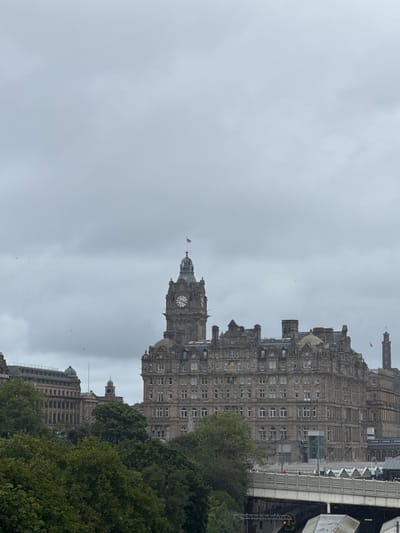 The image captures a view of the Balmoral Hotel in Edinburgh, Scotland, under a cloudy sky. The prominent clock tower of the Balmoral Hotel stands tall, its intricate stonework and clock faces clearly visible. Adjacent to it, a grand, multi-story building with numerous windows and ornate architectural details fills the frame. In the foreground, lush green trees provide a natural contrast to the urban landscape. Below the buildings, a railway bridge with visible tracks and a passing train suggests the presence of a transportation hub. The scene is set in the city of Edinburgh, United Kingdom, known for its historic architecture and picturesque cityscape.
