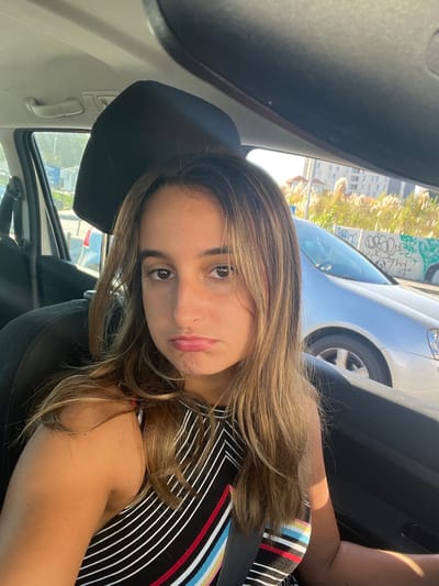 A young female, approximately 20-30 years old, is seated in the front passenger seat of a vehicle in Pedrouços, Portugal. She has long, light brown hair and is wearing a sleeveless top with diagonal stripes in black, white, red, yellow, and blue. Her lips are downturned, conveying a displeased or pouting expression. A dark seatbelt crosses her torso. Sunlight illuminates the scene from the right. Outside the vehicle, a silver sedan is visible on the right. Behind it, a low concrete wall is covered in extensive graffiti, primarily black and green. Beyond the wall, tall, feathery plants are prominent, with multi-story buildings visible in the background under a clear sky. On the left, a street scene with other parked vehicles and a white pole is partially visible through the driver's side window.