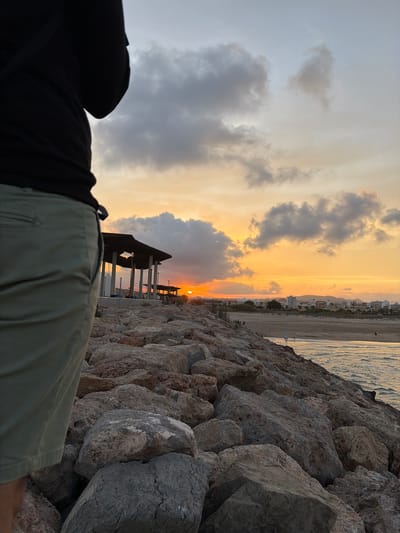 An observer stands on a rock breakwater at sunset in Sagunto, Spain. The foreground features the lower torso and legs of a person, seen from behind, wearing a dark top and olive green shorts, standing on large, irregular rocks. A dark strap from a bag or camera is visible over their left shoulder. The breakwater, composed of various grey, brown, and reddish rocks, extends diagonally into the midground, with gentle waves from the sea visible on the right. In the midground, a multi-columned, dark-roofed structure, possibly a pavilion or beach bar, is silhouetted against the setting sun. The sun, a bright orange disc, is visible low on the horizon, slightly to the right of this structure. The background reveals a wide sandy beach with faint human figures, and beyond it, a coastal town with multiple buildings against a backdrop of distant hills or mountains. The sky displays a dramatic sunset, transitioning from blue and grey overhead to vivid orange and yellow tones near the horizon, with scattered dark clouds illuminated by the sun's glow.