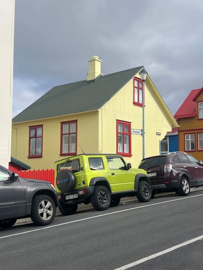 The image shows a street scene in Reykjavík, Iceland, at the coordinates 64.143 latitude and -21.923 longitude. A bright green Suzuki Jimny with a license plate reading "AL 279" is parked in the street, facing the camera. To its right, a maroon Chevrolet Suburban with a license plate reading "KP 78" is parked facing the same direction. The street is paved and lined with white lines. On the left side of the image, a gray SUV is partially obscured by a white building. Behind the vehicles, a yellow wooden house with red window frames stands on the corner of the street. The street sign on the house reads "Vitastíg". There is a green roof and a chimney on top of the house. The sky is overcast.