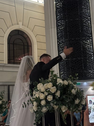 The image depicts an indoor event scene in Kiryat Ekron, Israel. A male figure, identified as a groom, is positioned to the right of a female figure, identified as a bride. The groom wears a black suit jacket over a white shirt with white cuffs, and has short, light-colored hair. His right arm is raised and extended to his right, with his hand open in a gesture. The bride, to the groom's left, wears a white wedding gown with a laced-up back and a long, sheer white veil that cascades down her back. Both figures are viewed from behind or the side.

In the lower foreground, a large, dense floral arrangement consisting of numerous white roses and green foliage, supported by a metallic stand, partially obscures the couple.

The background features ornate interior architecture. To the left, a light-colored wall displays recessed linear patterns and a prominent arched window with dark framing. To the right, a tall, dark decorative screen with intricate, repetitive leaf-like patterns is visible. A green illuminated sign is present in the upper right background. Partial views of additional individuals are visible in the background, out of focus, including a person in a turquoise shirt on the far left and a person in a light blue dress behind the floral arrangement on the right. In the lower right foreground, a hand holds a smartphone, its screen displaying an image of the event.