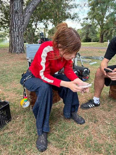 A person in a red jacket and blue jeans sits on outdoor equipment on a grassy surface, looking at a mobile phone. Another person in black shorts and shoes is partly visible from the waist up, holding a phone. A camera and a tree trunk are also visible.