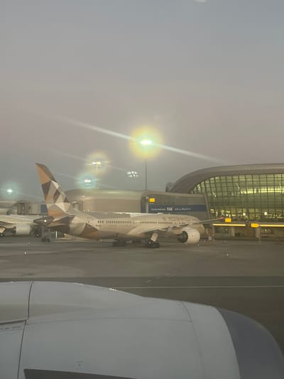 Two airplanes at an airport. One, an Etihad plane with a geometric brown and white pattern on the tail, is parked next to a terminal. Another, larger, white plane is partially visible on the right. Airport lighting is visible, creating a bright glare. The sky is overcast. The Etihad plane is parked adjacent to a boarding gate.