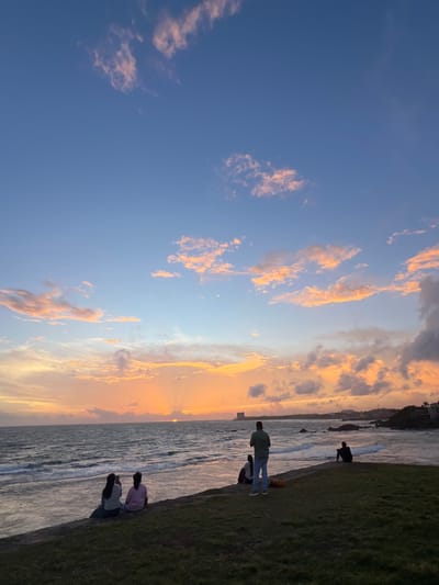 The image depicts a serene coastal scene at sunset in Galle, Sri Lanka. The sky is a canvas of blues and oranges, with scattered clouds catching the warm glow of the setting sun. The ocean stretches out to the horizon, its surface rippled by gentle waves. Along the shore, a grassy embankment meets a paved walkway.
Several individuals are present, enjoying the tranquil evening. In the foreground, a person in a green shirt and blue jeans stands facing the water, while another person sits beside them. Further along the walkway, two figures are seen sitting together, one in a pink shirt and the other in a light blue shirt. Another lone figure, silhouetted against the sky, is crouched by the water's edge. The atmosphere is peaceful and contemplative, suggesting a moment of relaxation and appreciation for the natural beauty.
The time of day is clearly sunset, as evidenced by the vibrant colors in the sky and the long shadows cast on the ground. The weather appears clear and pleasant, ideal for an evening stroll by the sea. The presence of the ocean, the distinctive clouds, and the general ambiance are strong cues for a coastal location. No visible text is present in the image.