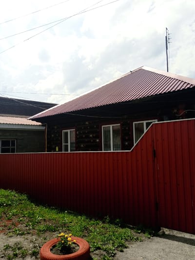 This media file shows a partially shaded view of a house with a dark wooden facade and a red corrugated metal roof. The house has several windows with white frames. A tall, dark red corrugated metal fence runs across the foreground, partially obscuring the house. To the left of the house, another building with a dark corrugated roof and a grey facade is visible. The sky above is partly cloudy. In the lower left corner, there is a patch of green grass and a red tire with yellow flowers planted inside. The scene appears to be outdoors during the daytime, with the sun casting shadows. The image evokes a sense of a residential setting, possibly a village or rural area, given the presence of the fence and the style of the house. No people are visible in the image. There is no discernible text in the image.