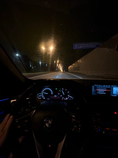 Captured from inside a vehicle, likely a BMW based on the steering wheel's emblem, the image shows a nighttime drive. Outside, streetlights illuminate a wet road, hinting at recent or ongoing rain. The vehicle's dashboard is visible, with the speedometer and infotainment system displaying data. The temperature reads 23.0 degrees Celsius. The presence of a hand on the steering wheel indicates a driver, although the person's features are obscured by the low light. Given the location, the scene is occurring in the city of Дубровка, Russia.