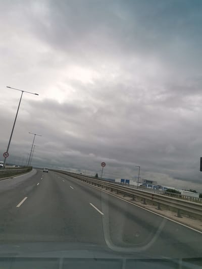The image depicts a highway with a gray, overcast sky. The highway has multiple lanes, with white lines marking the lanes and shoulders. There are streetlights along the side of the road. Two speed limit signs, marked with "80", are visible on the right side of the highway. In the distance, there are buildings and trees. A white car is driving on the highway, and other vehicles are visible in the distance. The scene is likely a view from inside a vehicle traveling on the highway. Based on the provided information, this highway segment is located in the city of Lozen, Bulgaria.