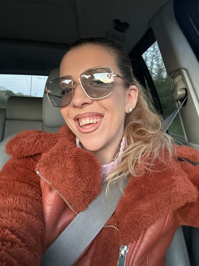 A woman is in a car, facing the camera at an angle, wearing a gray seatbelt, sunglasses, and a reddish-brown jacket with both furry and leather-like textures. She is smiling and exposing her teeth. There is a sunroof above. The location cannot be determined to be Grays, United Kingdom from the provided image.