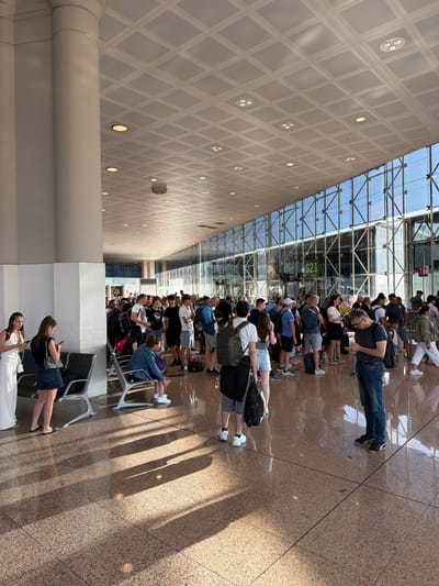 The image captures a scene inside an airport terminal, likely El Prat de Llobregat, Spain, given the context. A long line of people are standing, facing away from the camera, suggesting they are queuing for a flight, security, or check-in. The setting is modern and spacious, characterized by a high ceiling with a grid of diamond-shaped panels and recessed circular lights. A large glass wall or ceiling allows natural light to stream in, casting distinct shadows on the polished granite floor. The people are dressed casually, carrying luggage, including backpacks and suitcases. Some individuals are looking at their phones while waiting. The lighting suggests it is daytime, likely a bright and sunny day, as indicated by the strong shadows. The overall atmosphere appears to be one of patient waiting, common in travel hubs. Visible text includes "S21" on a sign behind some of the people, and "foodies" on a shopfront in the distance. The scene is predominantly viewed from a slightly elevated perspective, looking down the line of the queue.