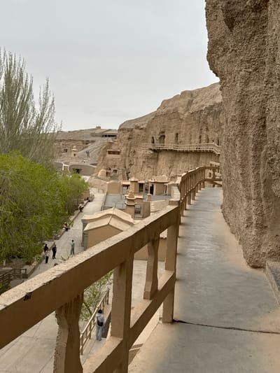 The image depicts a view of the Mogao Caves, located near Guazhou County, China. The caves are carved into a cliff face and feature a series of interconnected pathways and stairways. Several people are present, walking along the paths and exploring the site. The architectural style is characterized by its incorporation into the natural rock formations, with structures built directly into the cliff. Vegetation, including trees and shrubs, grows near the base of the cliff. The sky appears overcast. A stone railing or walkway structure is prominent in the foreground.