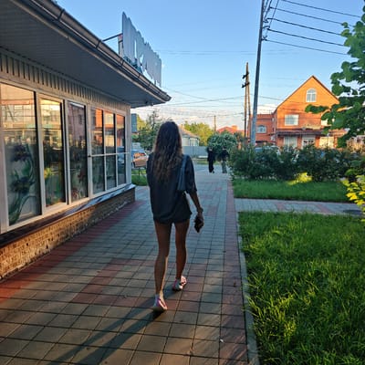 The media file shows a young woman with long, curly brown hair walking away from the camera on a paved sidewalk. She is wearing a dark gray t-shirt and light-colored shorts, with colorful athletic shoes. She is carrying a dark smartphone in her right hand and has a light gray shoulder bag slung over her left shoulder.
The scene appears to be a commercial street with a building on the left featuring large glass windows with floral stained-glass designs. The building has a metal awning and a sign with large, white, block letters, part of which is visible. The sidewalk is paved with red and gray rectangular tiles, and there is a strip of green grass next to it.
In the background, across the street, there is a two-story brick building with a prominent gable and arched windows. Further down the street, more buildings and trees are visible. Two people, one in black clothing and another in lighter attire, are walking on the sidewalk in the distance. Overhead, a network of power lines and utility poles are prominent against a clear blue sky.
The lighting suggests it is late afternoon or early evening, with long shadows cast by the building and the woman on the sidewalk. The sunlight highlights the grass, giving it a vibrant green hue. The overall atmosphere is calm and casual, depicting a person walking through a semi-urban environment on a pleasant day. Visible text on the building in the background appears to be in Russian, with one recognizable word being "шоколад" (chocolate).