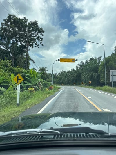 A view from inside a vehicle looking out at a road in Tambon Khlong Thom Nuea, Thailand. The road curves to the right, with a yellow line marking the center. On the left side of the road, there is a yellow diamond-shaped sign indicating an intersection. Further down the road, a yellow overhead sign displays Thai text and the number "60" inside a circle, suggesting a speed limit. Palm trees and dense green foliage line both sides of the road, with a cloudy sky overhead. Electrical poles and wires are visible. The vehicle's windshield and wipers are in the foreground.