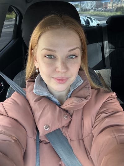 A young woman with light red hair and freckles is in the driver's seat of a car. She is wearing a light pink puffer jacket and a grey top. A grey seatbelt is fastened across her chest. She is looking at the camera with a slight smile. The car is parked, and a view of a building and street can be seen through the side window. The location is unknown.