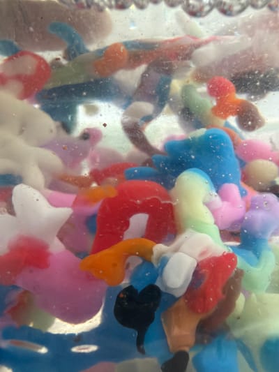 The image is a close-up, vertical shot of a collection of colorful, amorphous, semi-translucent objects submerged in a clear liquid, possibly water. The objects are predominantly soft, rounded shapes in various bright colors including red, blue, pink, orange, yellow, green, and white. Some of the objects appear to be shaped like small animals or gummy candies, such as bears or stars. Small air bubbles are dispersed throughout the liquid and cling to the surfaces of the objects, suggesting movement or agitation. The lighting appears to be coming from above and behind, creating highlights and some translucency. The background is indistinct and blurred, focusing all attention on the brightly colored objects. There is no discernible context for this scene in relation to the city of Папярнянскі сельскі Савет, Belarus, as the image depicts an abstract collection of objects without any identifying landmarks or features.