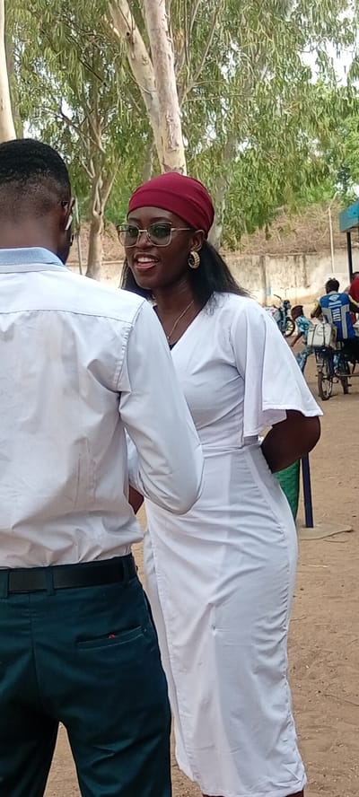 The image shows two people in an outdoor setting in Fagge D2, Nigeria. On the left, a man is seen from the back, wearing a white shirt and dark green pants with a black belt. He has short black hair and appears to be wearing glasses or earphones. To the right, a woman is facing him, wearing a white dress, a burgundy head covering, and gold hoop earrings. She has dark hair and is wearing glasses. Both seem to be engaged in a conversation. In the background, there are trees, a building, and other people, including one person riding a bicycle. The ground appears to be a dusty or sandy surface.