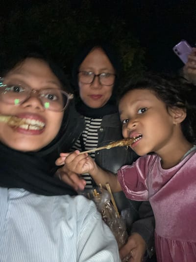 Three individuals, likely a family, are pictured in close proximity, engaging in eating from skewers at night. The foreground features a person on the left wearing glasses and a black hijab, in the process of biting into a skewer. Next to them, a younger child on the right, wearing a pink dress, is also eating from a skewer. In the background, a third individual, also wearing glasses and a hijab, is visible between the two in the foreground. The scene appears to be outdoors, with dark foliage suggesting a natural environment or a poorly lit urban area. The overall lighting is dim, consistent with nighttime. The presence of skewers and what appears to be casual clothing implies a relaxed, perhaps festive, occasion. Without further context, pinpointing the exact location within Tanggul, Indonesia, is not possible from the visual information alone.