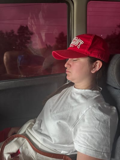 A young adult female is observed sleeping or resting within a vehicle located in Paje, Tanzania. She occupies the passenger side, framed from the mid-torso upward, with her head reclined against a grey fabric seat headrest. Her attire includes a red baseball cap with white embroidered text, partially legible as "ERGY UNIVERSITY," and a plain white short-sleeved t-shirt. A stud earring is visible in her left earlobe. Her eyes are closed, indicating a state of repose. A light-colored tote bag with brown leather handles rests in the lower left foreground, on her lap or seat. The vehicle's window, positioned to her left and behind her, is heavily tinted with a magenta hue. This tint obscures much of the external environment but clearly displays the reflection of an individual, likely the photographer, wearing light clothing and shorts/pants. Faint silhouettes of trees and overhead wires are discernible through the tinted glass, suggesting an outdoor, vegetated setting.