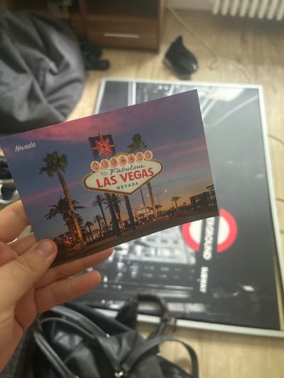 The image shows a hand holding a postcard featuring the iconic "Welcome to Fabulous Las Vegas Nevada" sign. The postcard is angled over a framed poster with a black and white design that includes a red circle with white lettering that appears to say "UNDERGROUND SUBWAY". The background includes a large grey beanbag chair and a wooden dresser or cabinet. There is also a black shoe and a black bag visible. The scene is indoors, with what appears to be a wooden floor. There is no indication of the city of Gelsenkirchen, Germany, in the image or postcard.