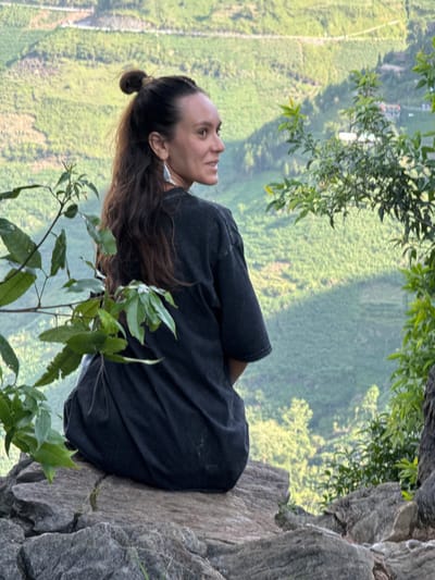 A woman with long brown hair pulled back in a bun sits on a rocky cliff overlooking a lush green valley. She is wearing a dark-colored t-shirt. The location is in Đồng Văn Commune, Vietnam, with terraced fields visible in the background. She is looking up and to her left. Branches with green leaves frame the foreground. The cliff edge is textured and gray.