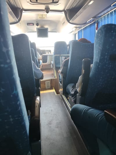 The image shows the interior of a bus, likely in transit. The seats are upholstered in blue fabric with a star-like pattern. Sunlight streams in through the windows, casting lens flares across the image. The aisle is visible, with wooden flooring. In the foreground, a person's legs are visible, wearing dark blue trousers. In the background, other passengers are seated, and a screen is mounted above the driver's area. Blue curtains are drawn along the windows on the right side. The image does not provide any specific visual cues to determine the location within the city of Zhanaarna, Kazakhstan.
