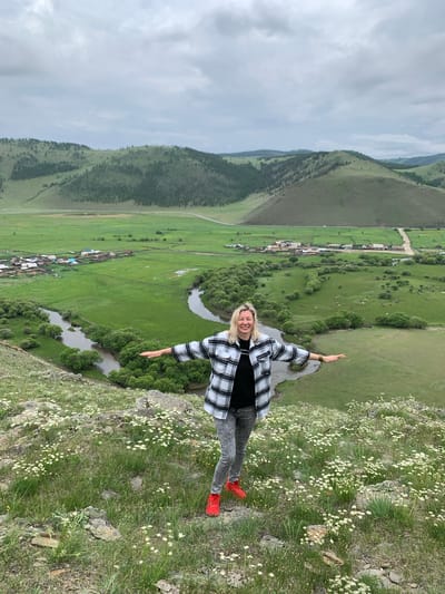 In Khurai-Nur, Russia, a woman stands on a grassy, rocky hill overlooking a valley. She is wearing a black t-shirt, a black and white checkered flannel shirt, distressed gray jeans, and bright red sneakers. Her arms are outstretched to her sides, and she is smiling. In the valley below, a winding river is visible, bordered by lush green trees. Further in the distance, a small village with wooden houses is situated in a vast green field. The landscape is characterized by rolling, tree-dotted hills under a cloudy sky.