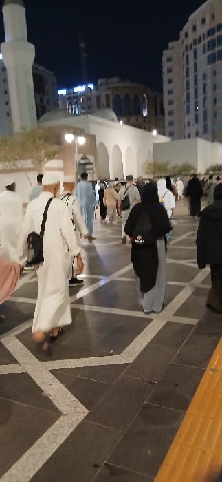 The image depicts a nighttime scene in an outdoor pedestrian plaza in Medina, Saudi Arabia, adjacent to the Prophet's Mosque (Masjid an-Nabawi). The foreground consists of dark grey square paving tiles interspersed with lighter grey-white geometric line patterns; a yellow tactile paving strip is visible on the far right. Numerous individuals, predominantly adults, are observed walking in various directions across the plaza. Many male subjects wear traditional white thobes and kufis, while several female subjects are clad in black abayas and hijabs.

In the left foreground, a person wears a light pink/peach garment. Immediately behind them, a male individual in a white thobe and white kufi walks away from the viewer, carrying a black shoulder bag and wearing black sandals. In the mid-ground, a female individual, seen from behind, wears a black abaya, grey pants, and a black backpack with an orange and black logo visible on its lower section.

The mid-ground architecture includes a low, white building with multiple arched openings and at least two domes, illuminated by artificial lighting. Streetlights with multiple globes are positioned within this area, alongside sparse, small trees or bushes. The background features a tall, slender white minaret with distinct architectural details, characteristic of the Prophet's Mosque. Adjacent to the minaret and extending across the background are several multi-story modern buildings, likely hotels or residential complexes, many of which are illuminated. A distant, partially obscured illuminated sign on one of these buildings is discernible. The overall environment is well-lit by artificial sources under a dark night sky.