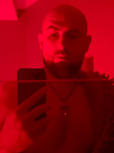 This is a selfie taken by a bald man with a beard, illuminated by red light. He is holding a dark-colored smartphone in his left hand, angled towards the camera, and appears to be looking at its screen. He is wearing a necklace with a cross pendant. The background is red and appears to be an interior space with a wall and possibly a ceiling light fixture visible in the upper left corner. There is a fuzzy, dark texture to the right of the man, which could be clothing or a decorative item. The image is grainy and underexposed due to the low light conditions and red illumination, obscuring fine details of the environment. There is no specific information in the image that allows for the identification of a location in Gliwice, Poland.